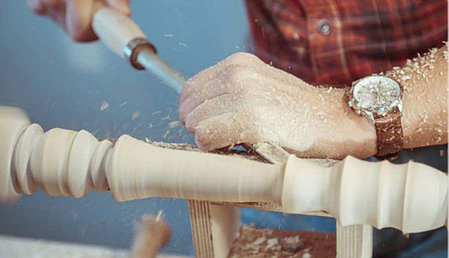 "Man wearing a red shirt and a watch, turning wood on a mini wood lathe with a chisel in hand"