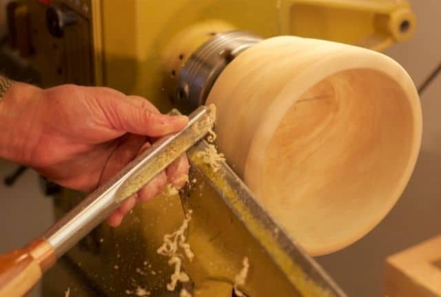 "Old man's hand holding a spindle gouge and turning a wooden bowl on a yellow colored lathe"